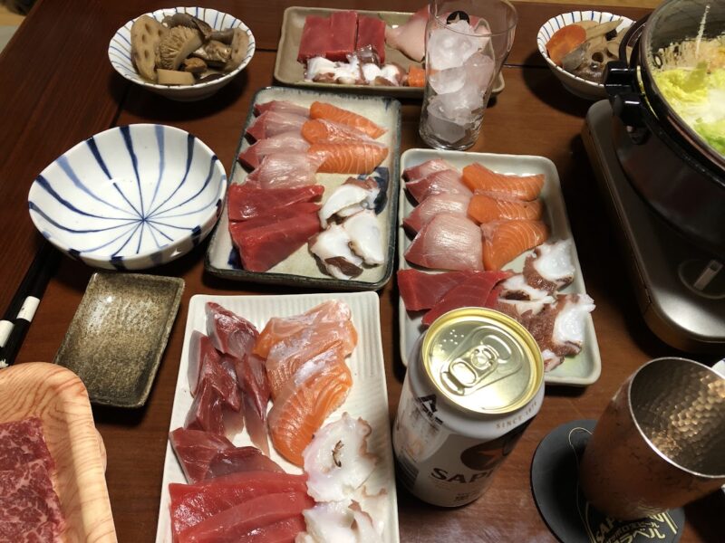 マグロ、サーモン、ハマチ、タコの刺身を複数の皿に盛り、缶ビールと並べた食卓