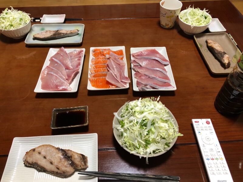 サーモンとブリの刺身、焼き魚、千切りキャベツのサラダが並んだ夕食の食卓