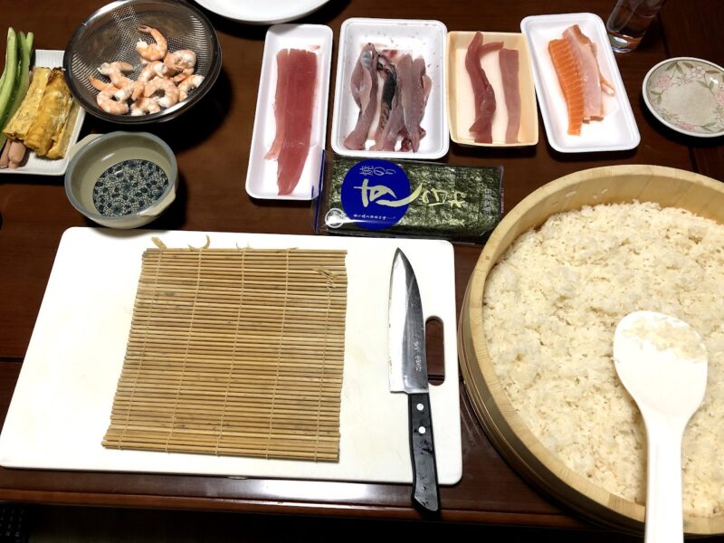 手巻き寿司用に切った刺身と玉子、エビ、海苔、寿司飯、包丁を並べた仕込みの様子